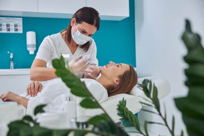 Brunette beautician in uniform holding syringe with an injection needle for Juvederm Voluma in Clinical Practice while patient lying on medicine chair in beauty salon