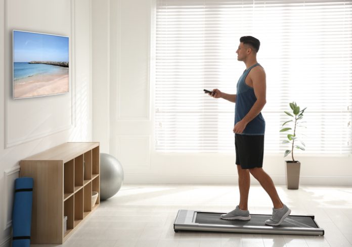 Sporty man with remote control training on walking treadmill and watching TV at home home workout space