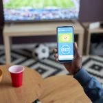 Close up of male hand holding smartphone with online sports bets on screen while watching football match at home,