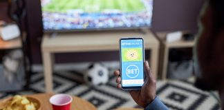 Close up of male hand holding smartphone with online sports bets on screen while watching football match at home,