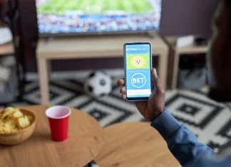 Close up of male hand holding smartphone with online sports bets on screen while watching football match at home,