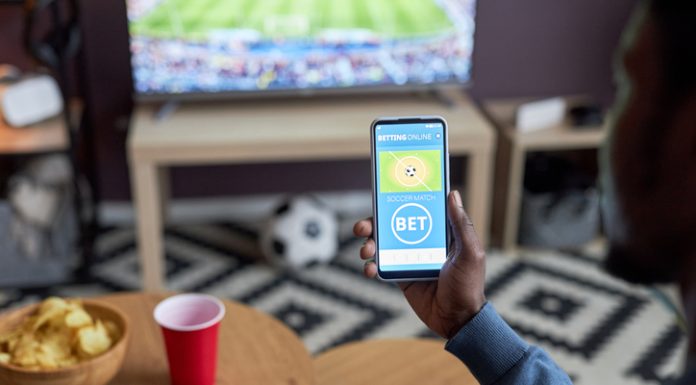 Close up of male hand holding smartphone with online sports bets on screen while watching football match at home,