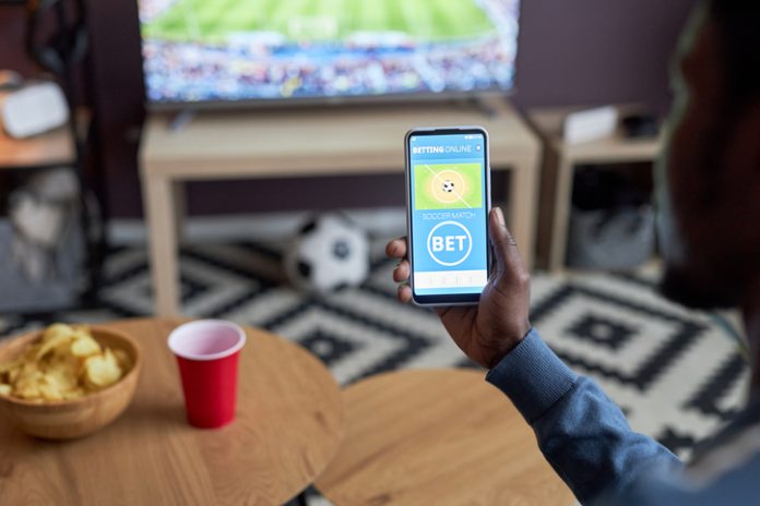 Close up of male hand holding smartphone with online sports bets on screen while watching football match at home,