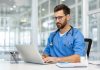Man doctor in blue scrubs works on laptop in bright, modern medical office. Doctor wearing stethoscope, using technology for research and communication. person health care