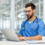 From Underdog to Innovator: Dr. Tomoaki Takei Journey Toward Whole Person Health Care Man doctor in blue scrubs works on laptop in bright, modern medical office. Doctor wearing stethoscope, using technology for research and communication. person health care