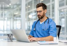 Man doctor in blue scrubs works on laptop in bright, modern medical office. Doctor wearing stethoscope, using technology for research and communication. person health care