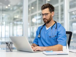 Man doctor in blue scrubs works on laptop in bright, modern medical office. Doctor wearing stethoscope, using technology for research and communication. person health care