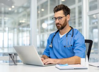 From Underdog to Innovator: Dr. Tomoaki Takei Journey Toward Whole Person Health Care Man doctor in blue scrubs works on laptop in bright, modern medical office. Doctor wearing stethoscope, using technology for research and communication. person health care