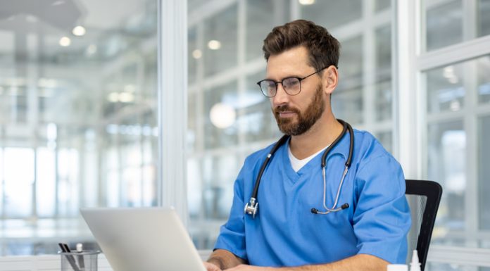 Man doctor in blue scrubs works on laptop in bright, modern medical office. Doctor wearing stethoscope, using technology for research and communication. person health care