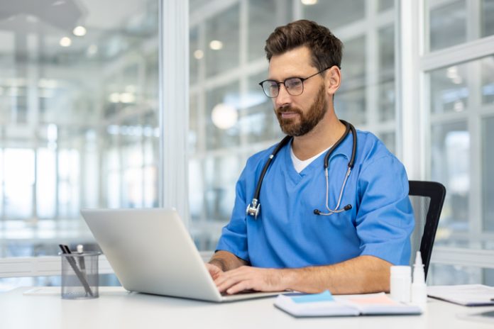 Male doctor in scrubs working on laptop in modern medical office. Professional medical worker with stethoscope using technology for research and communication. Healthcare and technology concept. Man doctor in blue scrubs works on laptop in bright, modern medical office. Doctor wearing stethoscope, using technology for research and communication. person health care