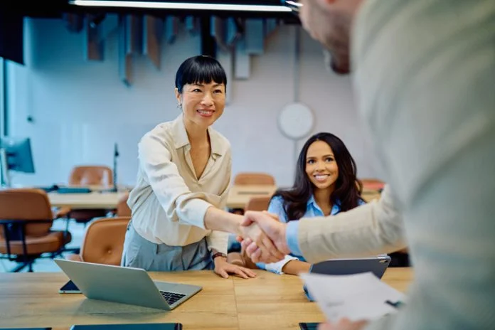 Diverse businesswomen greeting new team member with a handshake in a modern office, celebrating collaboration for Talent Strategy