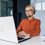 Top 5 Benefits of Integrating Virtual Receptionist Services into Your Business Model Successful businesswoman with video call headset working inside modern office, online customer support worker using laptop typing on keyboard, successful woman smiling satisfied with work.