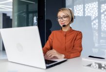 Successful businesswoman with video call headset working inside modern office, online customer support worker using laptop typing on keyboard, successful woman smiling satisfied with work.