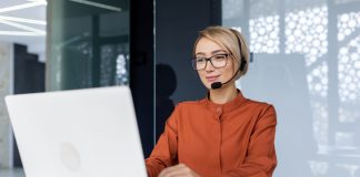 Successful businesswoman with video call headset working inside modern office, online customer support worker using laptop typing on keyboard, successful woman smiling satisfied with work.