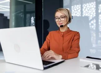 Successful businesswoman with video call headset working inside modern office, online customer support worker using laptop typing on keyboard, successful woman smiling satisfied with work.