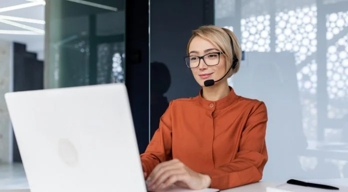 Successful businesswoman with video call headset working inside modern office, online customer support worker using laptop typing on keyboard, successful woman smiling satisfied with work.