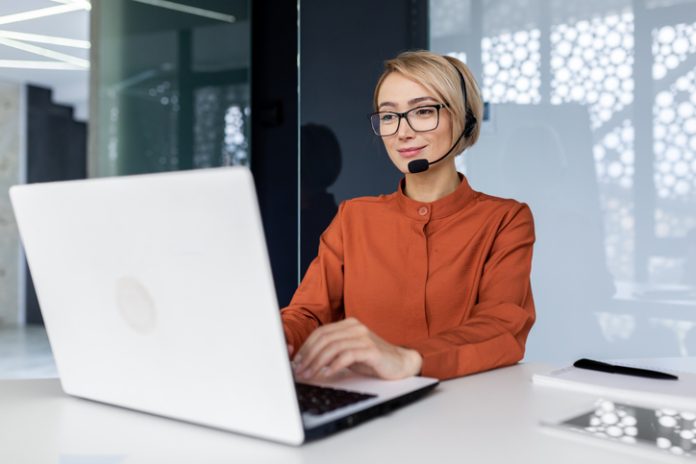 Successful businesswoman with video call headset working inside modern office, online customer support worker using laptop typing on keyboard, successful woman smiling satisfied with work.