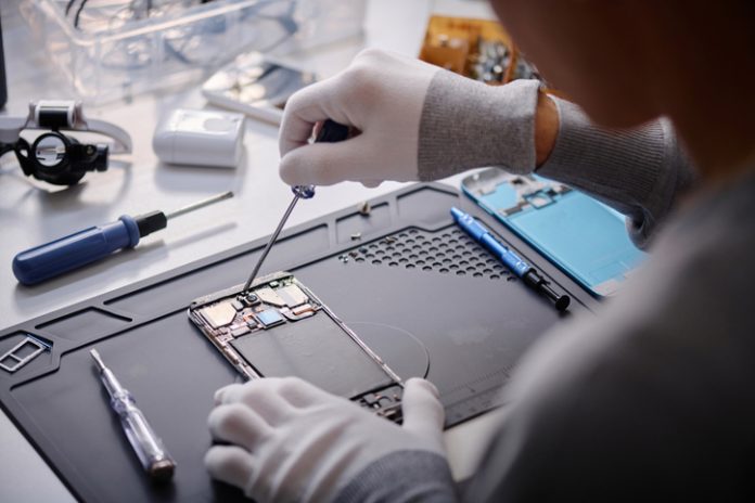Professional Repairman Working on Broken Cellphone at His Desk in Workshop Over shoulder shot of unrecognizable technician repairing disassembled smartphone, circuit with screwdriver