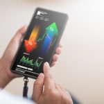 A close-up shot shows a person's hand holding a smartphone as tool, displaying a complex financial analysis interface with upward and downward trending percentage arrows, stock market charts, and various data points.