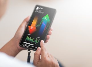 Prediction Markets are Becoming a Serious Business Tool: Here is What Executives Need to Know A close-up shot shows a person's hand holding a smartphone as tool, displaying a complex financial analysis interface with upward and downward trending percentage arrows, stock market charts, and various data points.