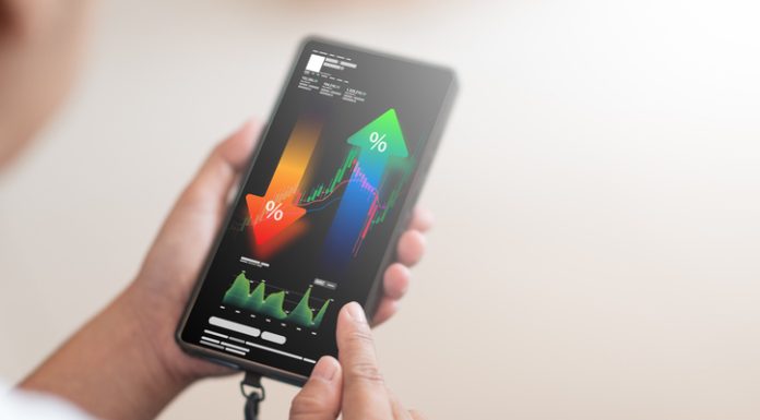 A close-up shot shows a person's hand holding a smartphone as tool, displaying a complex financial analysis interface with upward and downward trending percentage arrows, stock market charts, and various data points.