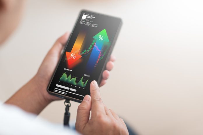 A close-up shot shows a person's hand holding a smartphone as tool, displaying a complex financial analysis interface with upward and downward trending percentage arrows, stock market charts, and various data points.