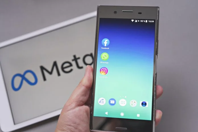 Mobile display with logo of Facebook, WhatsApp and Instagram apps in against blurred META logotype