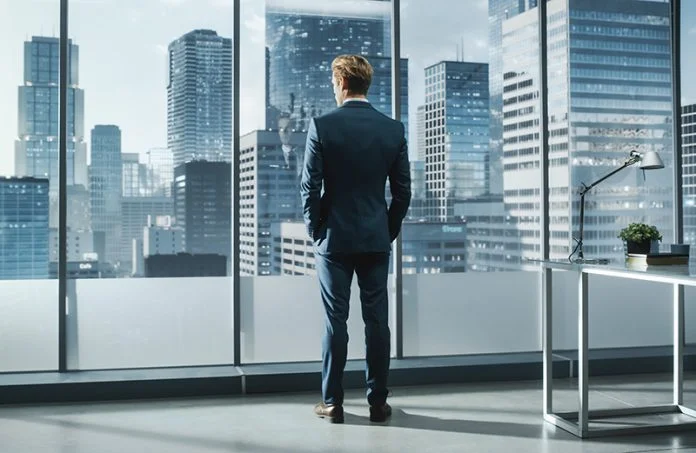 Confident Young Businessman in a Perfect Tailored Suit Standing in His Modern Office Looking out of the Window on Big City with Skyscrapers. Successful Finance Manager Planning Project Strategy. CEO Tenure, Succession, and the Value of the Firm