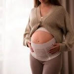 How a Pregnancy Belt Can Improve Posture and Mobility for Active Moms-to-Be Pregnant woman wearing comfortable, orthopedic belly belt, bandage, touching baby bump near window
