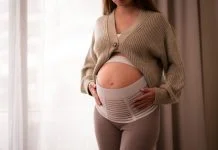 How a Pregnancy Belt Can Improve Posture and Mobility for Active Moms-to-Be Pregnant woman wearing comfortable, orthopedic belly belt, bandage, touching baby bump near window