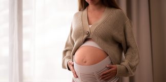 How a Pregnancy Belt Can Improve Posture and Mobility for Active Moms-to-Be Pregnant woman wearing comfortable, orthopedic belly belt, bandage, touching baby bump near window