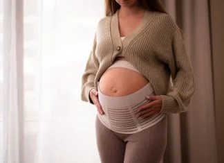 Pregnant woman wearing comfortable, orthopedic belly belt, bandage, touching baby bump near window