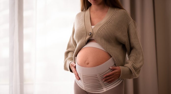 How a Pregnancy Belt Can Improve Posture and Mobility for Active Moms-to-Be Pregnant woman wearing comfortable, orthopedic belly belt, bandage, touching baby bump near window
