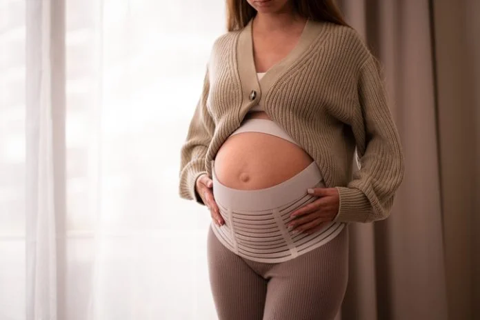 Pregnant woman wearing comfortable, orthopedic belly belt, bandage, touching baby bump near window