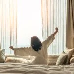 Asian woman waking up from good sleep on bed in weekend morning resting in comfort bedroom looking toward city view, having happy, productivity, work-life quality balance lifestyle