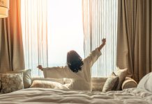 Asian woman waking up from good sleep on bed in weekend morning resting in comfort bedroom looking toward city view, having happy, productivity, work-life quality balance lifestyle