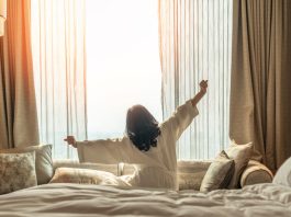 How Productivity Improves Based on Where You Wake Up Asian woman waking up from good sleep on bed in weekend morning resting in comfort bedroom looking toward city view, having happy, productivity, work-life quality balance lifestyle