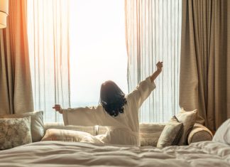 Asian woman waking up from good sleep on bed in weekend morning resting in comfort bedroom looking toward city view, having happy, productivity, work-life quality balance lifestyle