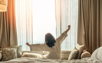 Asian woman waking up from good sleep on bed in weekend morning resting in comfort bedroom looking toward city view, having happy, productivity, work-life quality balance lifestyle