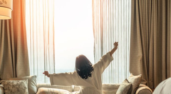 How Productivity Improves Based on Where You Wake Up Asian woman waking up from good sleep on bed in weekend morning resting in comfort bedroom looking toward city view, having happy, productivity, work-life quality balance lifestyle