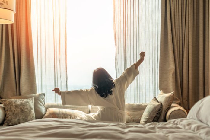 Asian woman waking up from good sleep on bed in weekend morning resting in comfort bedroom looking toward city view, having happy, productivity, work-life quality balance lifestyle