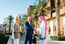 Mature Arabic businessman shaking hands with Caucasian partners in Jeddah Culture, Saudi Arabia Square featuring traditional architecture and palm trees.