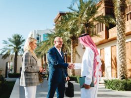 Mature Arabic businessman shaking hands with Caucasian partners in Jeddah Culture, Saudi Arabia Square featuring traditional architecture and palm trees.
