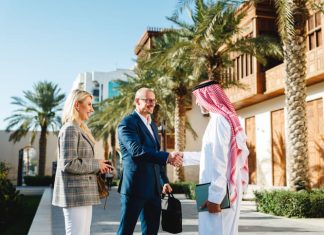 Mature Arabic businessman shaking hands with Caucasian partners in Jeddah Culture, Saudi Arabia Square featuring traditional architecture and palm trees.