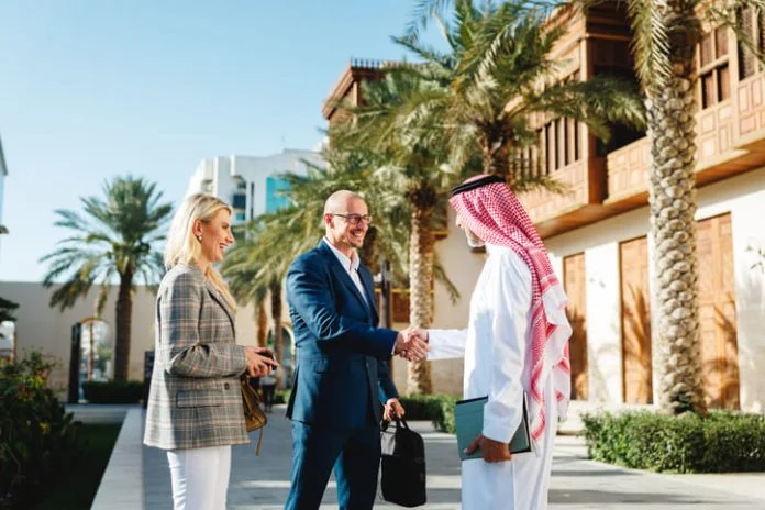 iStock-2238711897 (1) Mature Arabic businessman shaking hands with Caucasian partners in Jeddah Culture, Saudi Arabia Square featuring traditional architecture and palm trees.