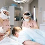 Two female trichologist performing local needle-free anesthesia to back of head using pistol in recipient area prior to hair transplant surgery in clinic.