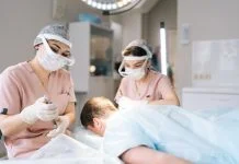 Two female trichologist performing local needle-free anesthesia to back of head using pistol in recipient area prior to hair transplant surgery in clinic.