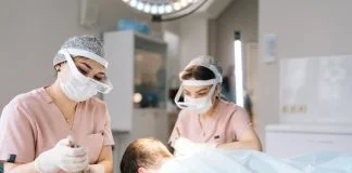Two female trichologist performing local needle-free anesthesia to back of head using pistol in recipient area prior to hair transplant surgery in clinic.