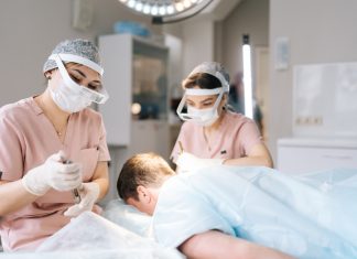 Two female trichologist performing local needle-free anesthesia to back of head using pistol in recipient area prior to hair transplant surgery in clinic.