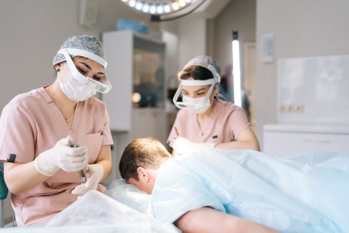 Two female trichologist performing local needle-free anesthesia to back of head using pistol in recipient area prior to hair transplant surgery in clinic.